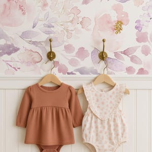 May include: Two baby outfits hanging on wooden hangers against a floral wallpaper backdrop. One is a rust-colored long-sleeved bodysuit dress. The other is a white romper with a pink floral pattern and ruffled trim.