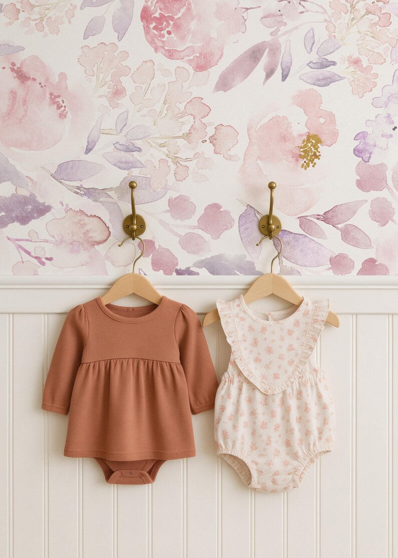 May include: Two baby outfits hanging on wooden hangers against a floral wallpaper backdrop. One is a rust-colored long-sleeved bodysuit dress. The other is a white romper with a pink floral pattern and ruffled trim.