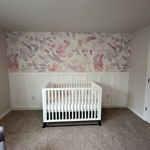 May include: A white crib with a mattress sits in a room with a floral wallpaper backdrop. The wallpaper features pink and purple flowers and leaves. The room has white wainscoting and a neutral-colored carpet.