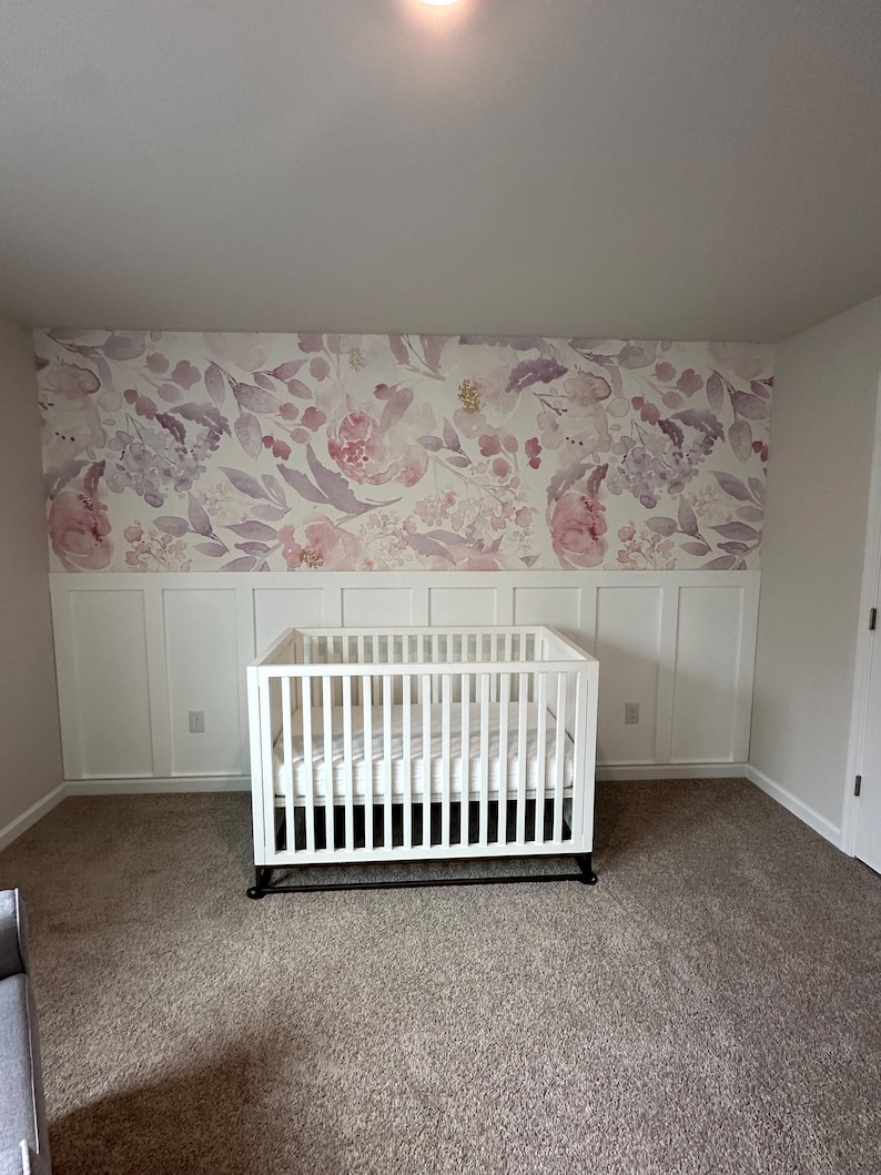 May include: A white crib with a mattress sits in a room with a floral wallpaper backdrop. The wallpaper features pink and purple flowers and leaves. The room has white wainscoting and a neutral-colored carpet.
