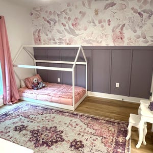 May include: A child's bedroom with a white house-shaped bed frame, a pink comforter, and a floral patterned wallpaper. A large area rug with a distressed pattern in shades of pink, purple, and cream covers the floor. A white ceiling fan is visible.