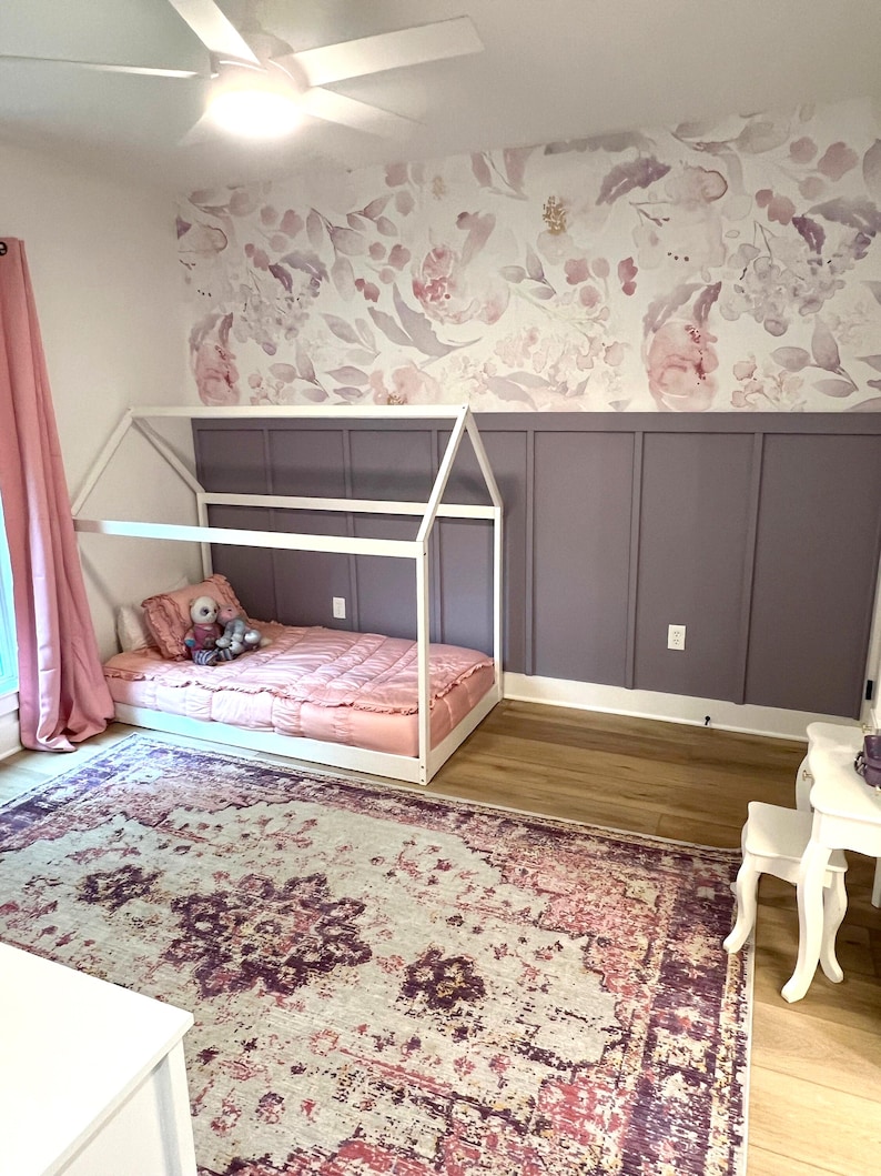 May include: A child's bedroom with a white house-shaped bed frame, a pink comforter, and a floral patterned wallpaper. A large area rug with a distressed pattern in shades of pink, purple, and cream covers the floor. A white ceiling fan is visible.