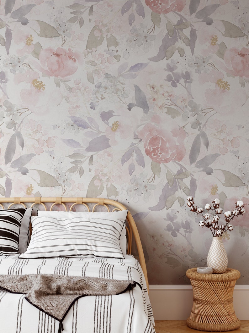 May include: A bedroom scene featuring a floral wallpaper with pink and purple watercolor flowers. A bed with striped bedding and pillows is in the foreground, along with a woven headboard and a small side table with a vase of cotton stems.