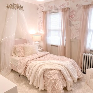 May include: A light pink bedroom with a bed featuring a floral-patterned comforter and a fluffy white throw blanket. The room includes sheer pink curtains, a white rug, and a decorative canopy with fairy lights, creating a soft and inviting atmosphere.