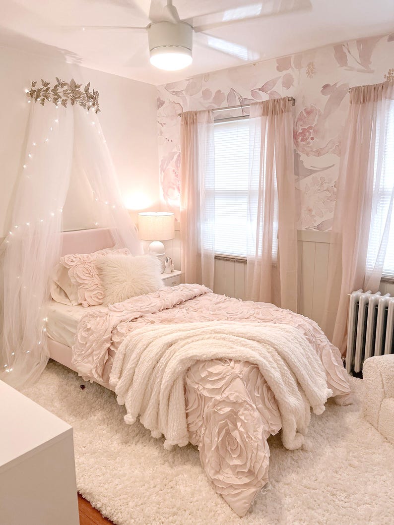 May include: A light pink bedroom with a bed featuring a floral-patterned comforter and a fluffy white throw blanket. The room includes sheer pink curtains, a white rug, and a decorative canopy with fairy lights, creating a soft and inviting atmosphere.