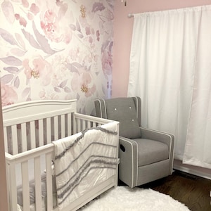 May include: A nursery with a white crib, a gray rocking chair, and a fluffy white rug. The wall features a floral wallpaper in shades of pink and purple. White curtains hang on the right side of the room.
