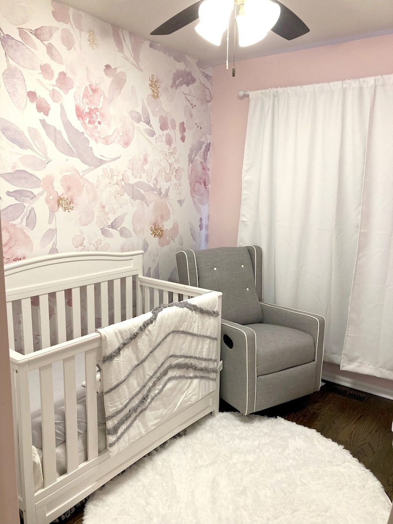 May include: A nursery with a white crib, a gray rocking chair, and a fluffy white rug. The wall features a floral wallpaper in shades of pink and purple. White curtains hang on the right side of the room.