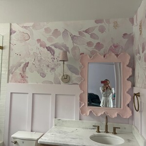 May include: A bathroom with a floral wallpaper in shades of pink and purple. A light pink, wavy-edged mirror hangs above a marble countertop with a white sink and gold faucet. A matching pink wall paneling is below the wallpaper.