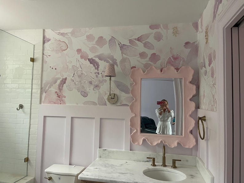 May include: A bathroom with a floral wallpaper in shades of pink and purple. A light pink, wavy-edged mirror hangs above a marble countertop with a white sink and gold faucet. A matching pink wall paneling is below the wallpaper.