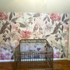 May include: A gold metal crib in a room with floral wallpaper. The wallpaper features large pink and purple flowers with green leaves on a white background. The crib is centered in front of the wall.