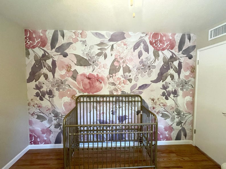 May include: A gold metal crib in a room with floral wallpaper. The wallpaper features large pink and purple flowers with green leaves on a white background. The crib is centered in front of the wall.