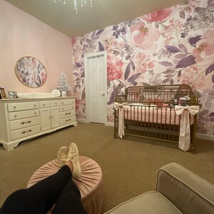 May include: A nursery with a floral wallpaper in shades of pink and purple. A white dresser, a crib with pink bedding, and a pink ottoman are visible. A chandelier hangs from the ceiling.
