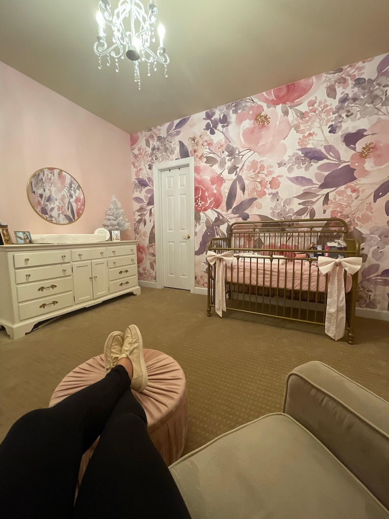May include: A nursery with a floral wallpaper in shades of pink and purple. A white dresser, a crib with pink bedding, and a pink ottoman are visible. A chandelier hangs from the ceiling.