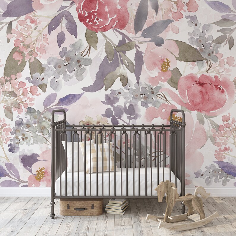 May include: A nursery scene featuring a wrought iron crib with a white mattress and a plaid pillow. The backdrop is a floral watercolor wallpaper in shades of pink, purple, and gray. A wooden rocking horse and a small suitcase are also present.