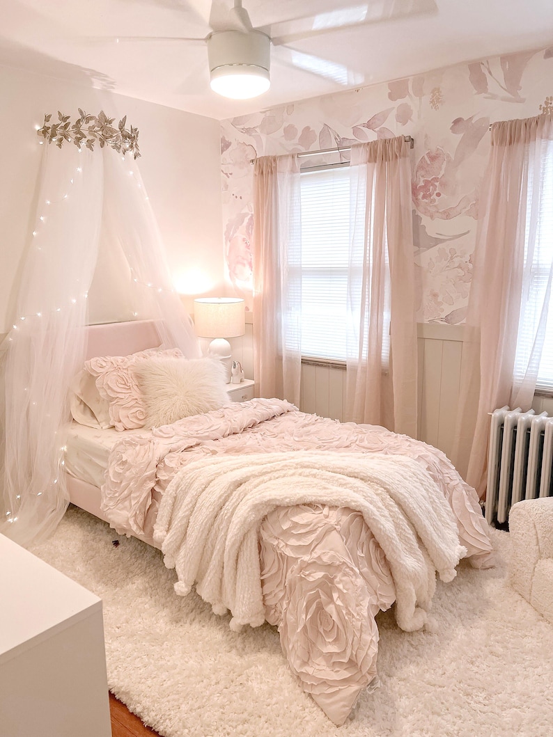 May include: A light pink bedroom with a bed featuring a floral-patterned comforter and a fluffy white throw blanket. The room includes sheer pink curtains, a floral wallpaper, and a white rug. A canopy with fairy lights hangs above the bed.