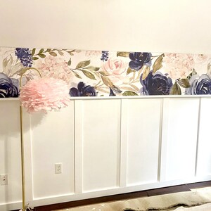 May include: A floral wallpaper border featuring watercolor-style navy and blush roses and greenery adorns the wall above a white wainscoting. A gold floor lamp with a pink feather shade stands in front of the wall.
