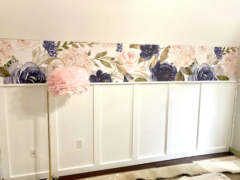 May include: A floral wallpaper border featuring watercolor-style navy and blush roses and greenery adorns the wall above a white wainscoting. A gold floor lamp with a pink feather shade stands in front of the wall.