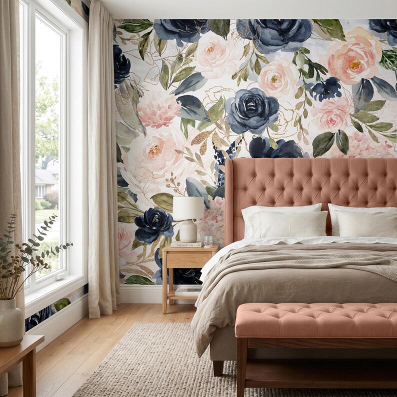 May include: A bedroom featuring a floral wallpaper mural with navy blue and blush pink roses, green leaves, and gold accents. A tufted salmon-colored headboard and matching bench sit in front of the wall, paired with white bedding and a beige throw.