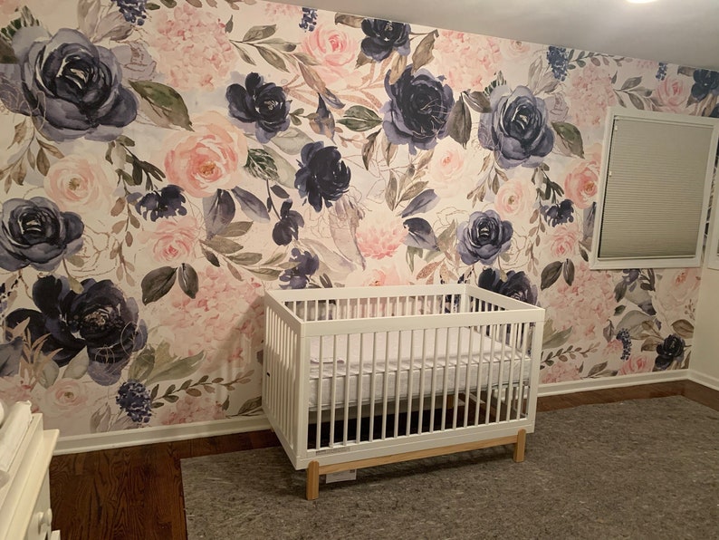 May include: A white wooden crib with a mattress sits against a wall covered in floral wallpaper featuring large navy blue and soft pink roses, green leaves, and delicate stems. The wallpaper pattern covers the entire wall behind the crib.
