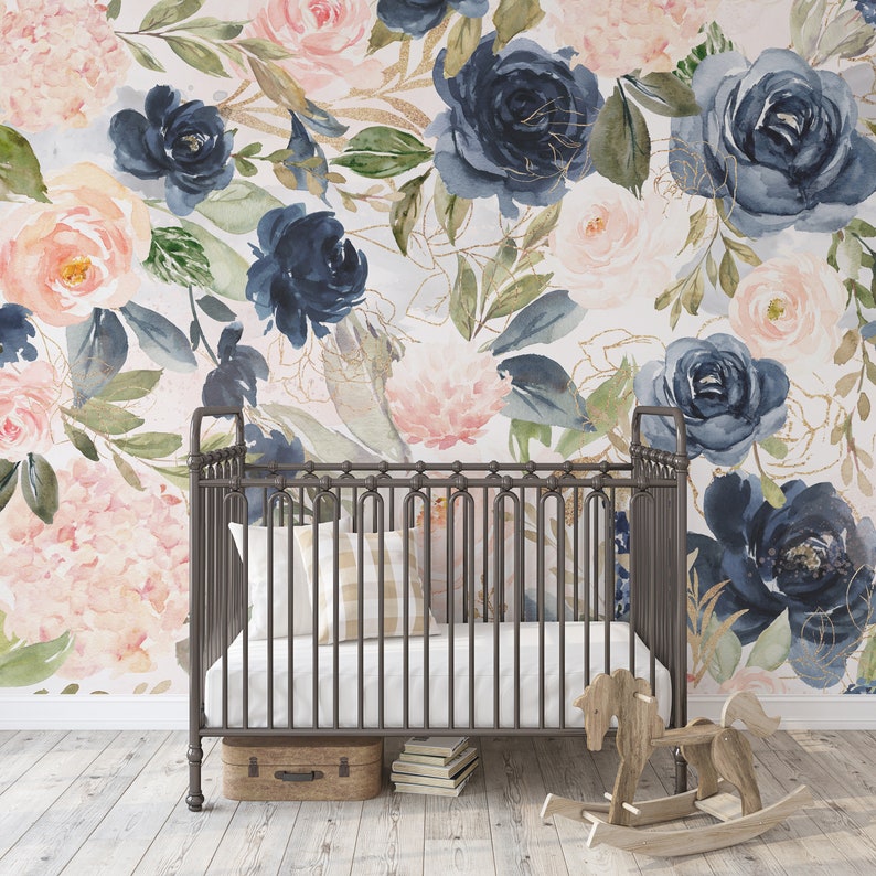 May include: A nursery scene featuring a wrought iron crib with a white mattress and a plaid pillow. The backdrop is a floral wallpaper with navy, blush, and gold accents. A wooden rocking horse and a small suitcase are in the foreground.