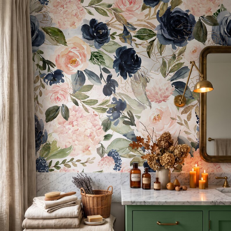 May include: A bathroom vanity area featuring floral wallpaper with navy blue and blush pink roses, green leaves, and gold accents. The setup includes a green cabinet, a marble countertop, a wicker basket, stacked towels, and lit candles.