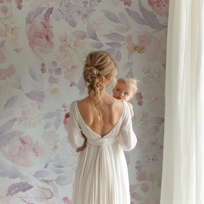 May include: A woman in a white dress holds a baby in front of a floral wallpaper. The wallpaper features watercolor-style flowers in shades of pink and purple. A sheer white curtain is on the right side of the image.