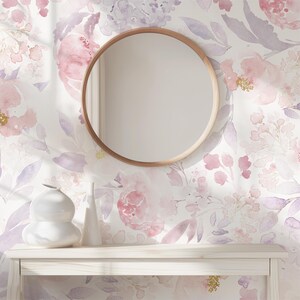 May include: A round wooden-framed mirror hangs on a wall with a floral watercolor design in pink and purple hues. Below the mirror, a white table displays two white ceramic vases. The overall aesthetic is soft and feminine.