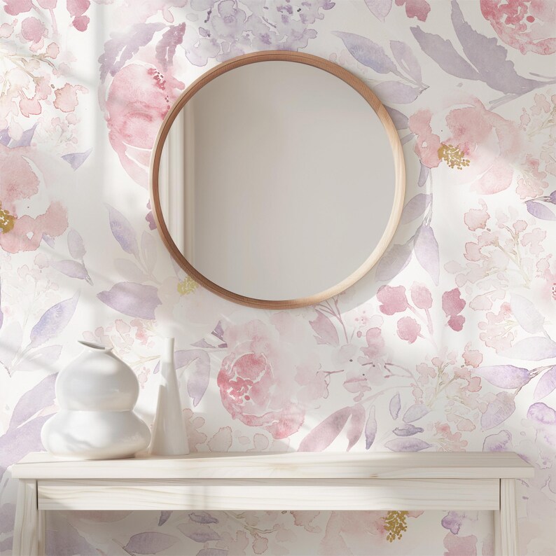 May include: A round wooden-framed mirror hangs on a wall with a floral watercolor design in pink and purple hues. Below the mirror, a white table displays two white ceramic vases. The overall aesthetic is soft and feminine.