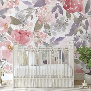 May include: A white crib with a striped skirt and pillows sits in front of a floral watercolor wallpaper. The wallpaper features large pink and purple flowers with gray and green leaves. A wooden rocking horse and a potted plant are in the foreground.