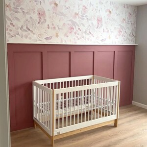 May include: A white crib with a light wood base sits in a nursery. The walls are painted a deep red with white trim and a floral wallpaper. The floor is light wood.