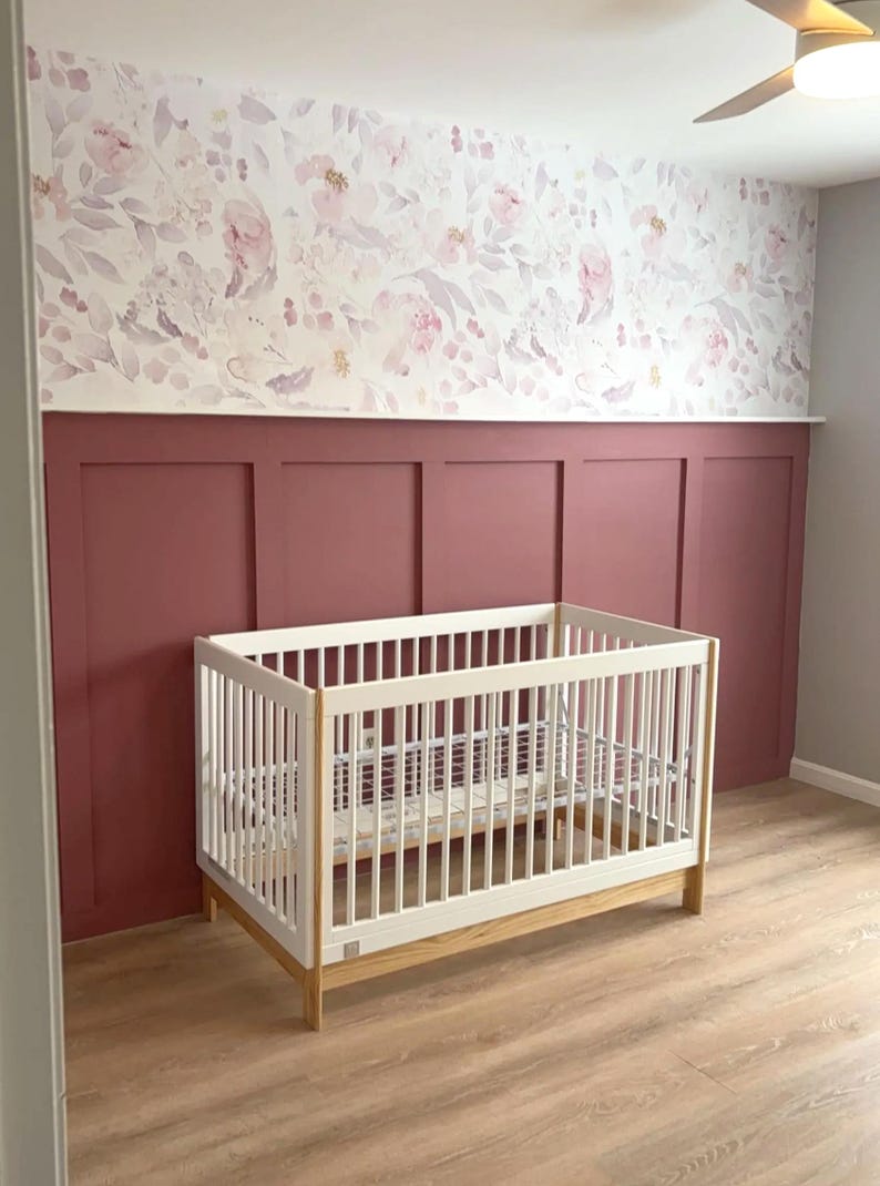 May include: A white crib with a light wood base sits in a nursery. The walls are painted a deep red with white trim and a floral wallpaper. The floor is light wood.