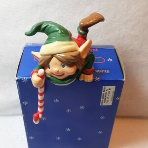 May include: A Christmas stocking holder featuring a green and red elf with brown hair and blue eyes. The elf is holding a candy cane and peeking over the edge of a blue box with snowflakes. The box reads "Ocean Desert Sales Hammonton, NJ Item No. X'mas 67 Made in China".