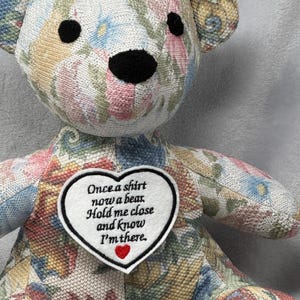 May include: A stuffed bear made from floral patterned fabric with black button eyes and a black nose. A white heart-shaped patch on the chest features black embroidered text that reads: Once a shirt now a bear. Hold me close and know I'm there.