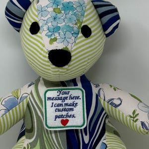 May include: A stuffed bear constructed from various fabric patches in shades of green, blue, and white. The bear features a floral pattern, stripes, and a white square patch on its chest with the text: Your message here. I can make custom patches.