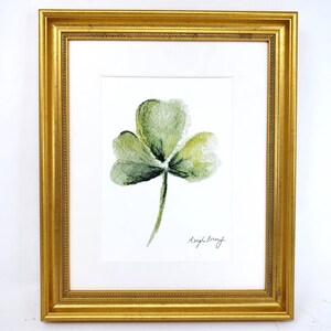 May include: A framed watercolor painting of a green three-leaf clover. The artwork is set against a white background and framed in a gold-colored, ornate frame. The artist's signature is visible in the lower right corner.