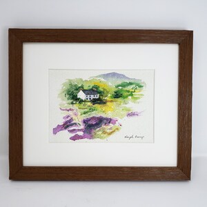 May include: Watercolour painting of a white cottage with a dark roof, set in a vibrant landscape of green, yellow, and purple hues. The artwork is framed in a brown wooden frame with a white mount.