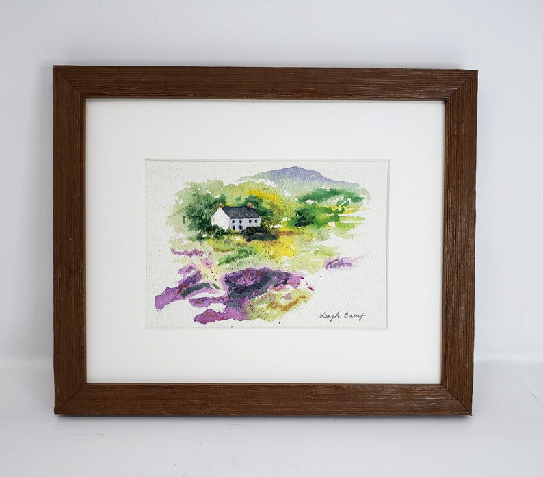 May include: Watercolour painting of a white cottage with a dark roof, set in a vibrant landscape of green, yellow, and purple hues. The artwork is framed in a brown wooden frame with a white mount.