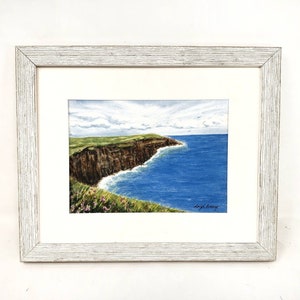 May include: A framed watercolor painting depicts a coastal landscape. The artwork showcases a brown cliff, green grass, and blue ocean under a cloudy sky. The painting is signed and framed in a white wooden frame.