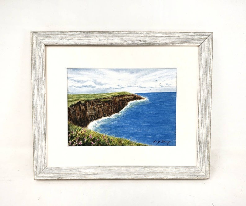 May include: A framed watercolor painting depicts a coastal landscape. The artwork showcases a brown cliff, green grass, and blue ocean under a cloudy sky. The painting is signed and framed in a white wooden frame.