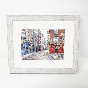 May include: Framed watercolour painting of a European street scene. The artwork shows a cobbled street with buildings in blue, brown, and red. A pub with the text "The Temple Bar" is visible. The frame is white wood.