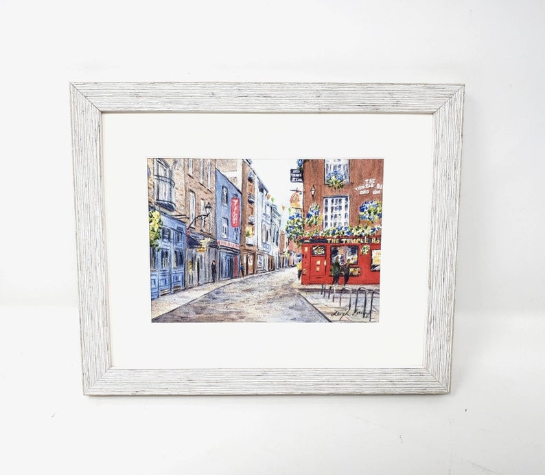 May include: Framed watercolour painting of a European street scene. The artwork shows a cobbled street with buildings in blue, brown, and red. A pub with the text "The Temple Bar" is visible. The frame is white wood.