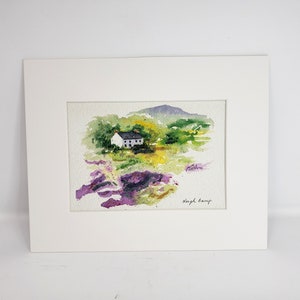 May include: Watercolour painting of a white house with a dark roof set in a green and yellow landscape, with a purple mountain in the background. The artwork is matted in a white frame. Signed by Leigh Barry.