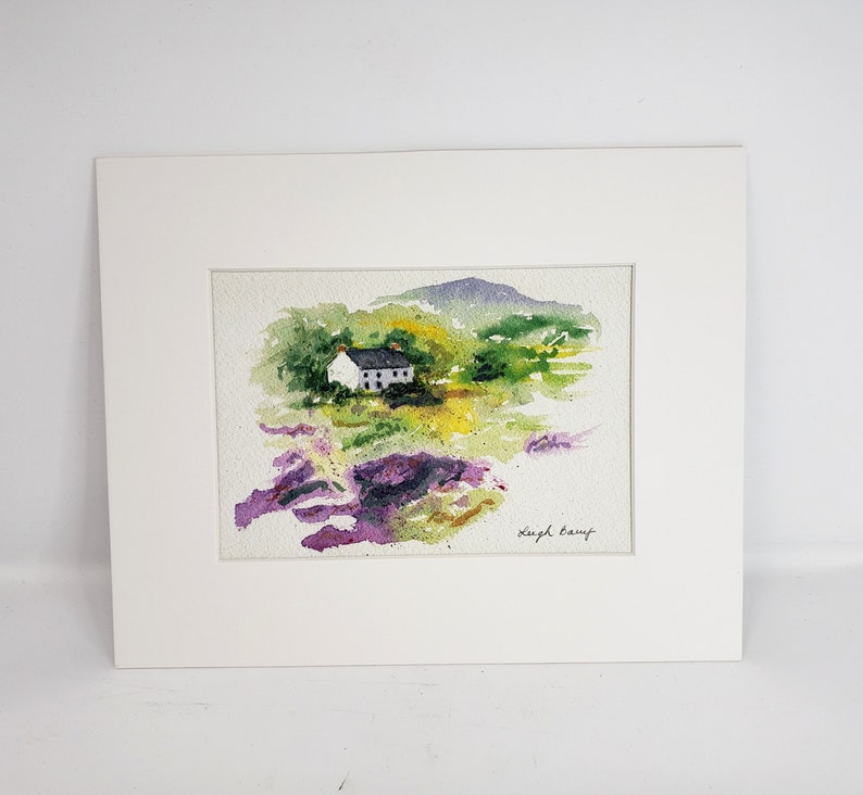 May include: Watercolour painting of a white house with a dark roof set in a green and yellow landscape, with a purple mountain in the background. The artwork is matted in a white frame. Signed by Leigh Barry.