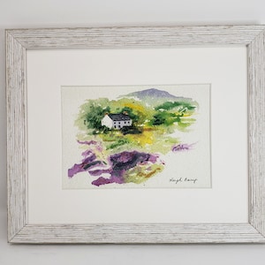 May include: Watercolour painting of a white cottage with a black roof in a green and yellow landscape, framed in a white distressed wood frame. Purple and pink flowers in the foreground. Signed by Leigh Lamp.