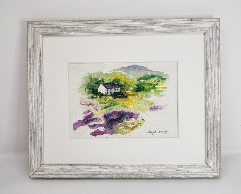 May include: Watercolour painting of a white cottage with a black roof in a green and yellow landscape, framed in a white distressed wood frame. Purple and pink flowers in the foreground. Signed by Leigh Lamp.
