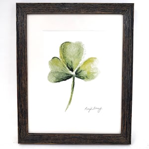 May include: Watercolor painting of a green shamrock with three leaves, signed "Leigh Barry", framed in a dark brown wooden frame. The shamrock is centered on a white background.