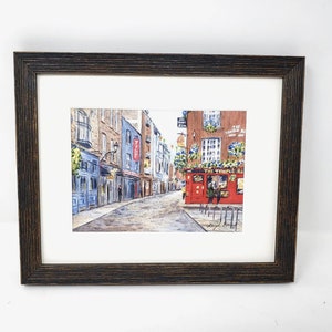May include: Framed watercolour painting of a European street scene. The artwork shows a cobblestone street with buildings, including a red pub with the text "The Temple Bar". The frame is dark brown wood.