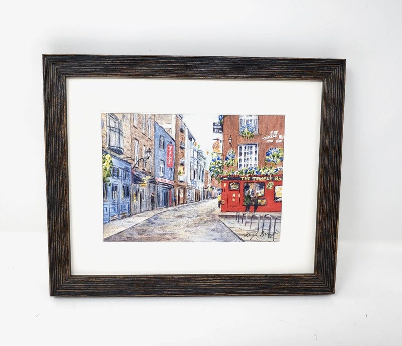 May include: Framed watercolour painting of a European street scene. The artwork shows a cobblestone street with buildings, including a red pub with the text "The Temple Bar". The frame is dark brown wood.
