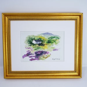 May include: Watercolour painting of a white cottage with a black roof, set in a green and purple landscape. The artwork is framed in a gold-coloured, ornate frame with a white mount.