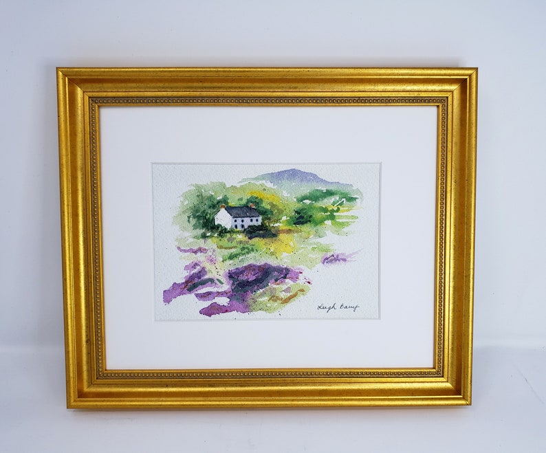 May include: Watercolour painting of a white cottage with a black roof, set in a green and purple landscape. The artwork is framed in a gold-coloured, ornate frame with a white mount.